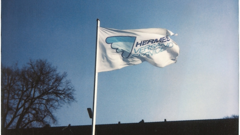 Ein Mast mit einer weißen Flagge ragt in den Himmel, darauf ist das blaue Hermes Logo aus den 1970er Jahren zu sehen (Foto: Hermes).
