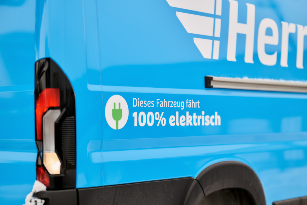 Ausschnitt eines Fotos von einem Hermes E-Transporter. Das Hermes-Logo ist angeschnitten und es ist der Schriftzug "Dieses Fahrzeug fährt zu 100% elektrisch" zu lesen.