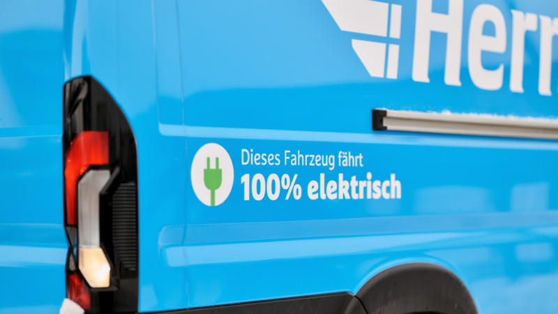 Ausschnitt eines Fotos von einem Hermes E-Transporter. Das Hermes-Logo ist angeschnitten und es ist der Schriftzug "Dieses Fahrzeug fährt zu 100% elektrisch" zu lesen.