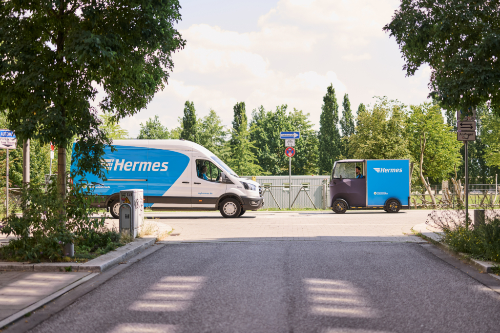 Hermes E-Transporter und Lastenrad fahren auf einer Straße auf gegenüberliegenden Seiten aneinander vorbei. Im Hintergrund ist ein Einbahnstraßenschild zu sehen mit dem ergänzenden Schild, dass Fahrräder in beide Richtungen fahren dürfen.